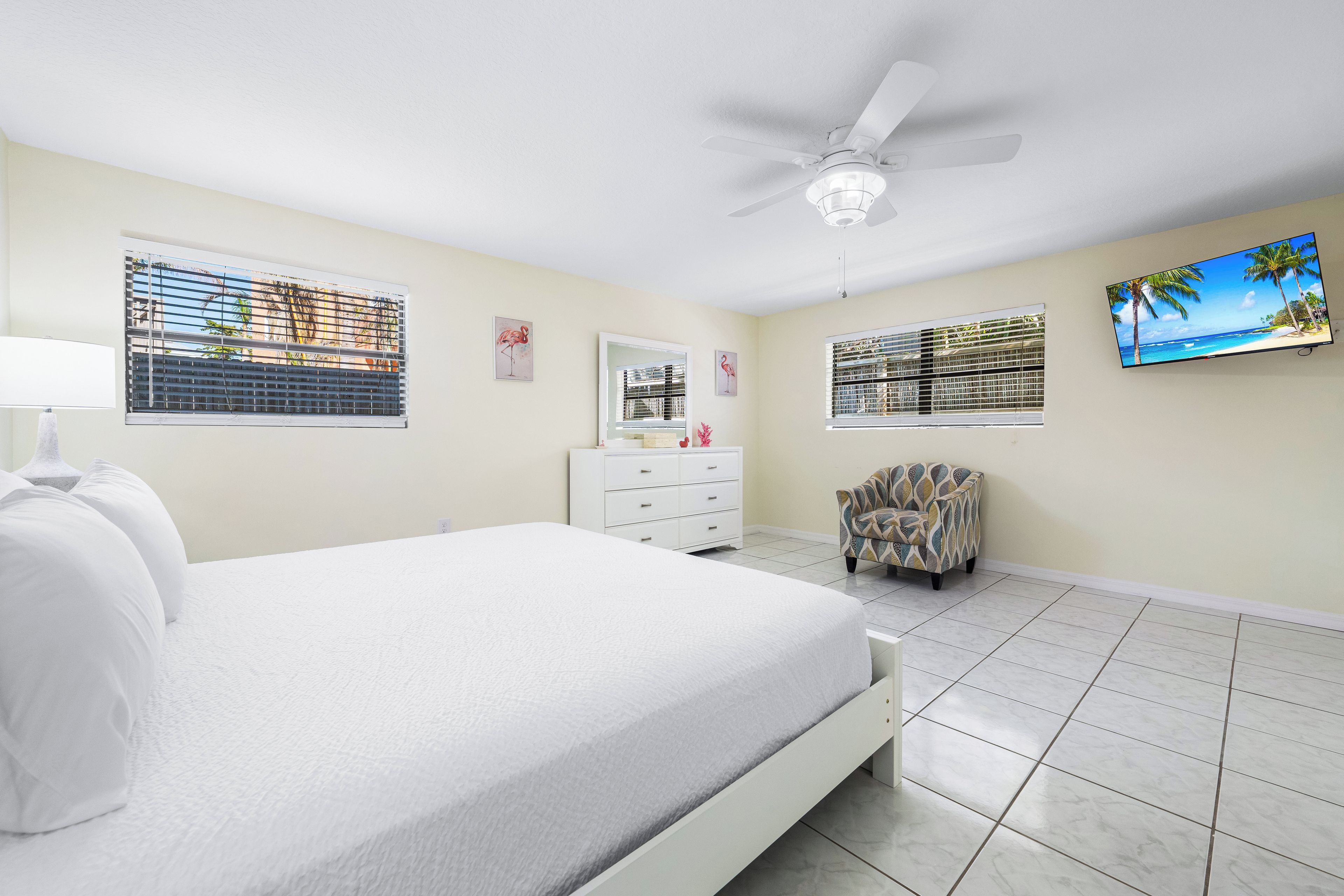 A bedroom with a bed, a dresser, a chair, a ceiling fan, a TV, and windows with blinds.