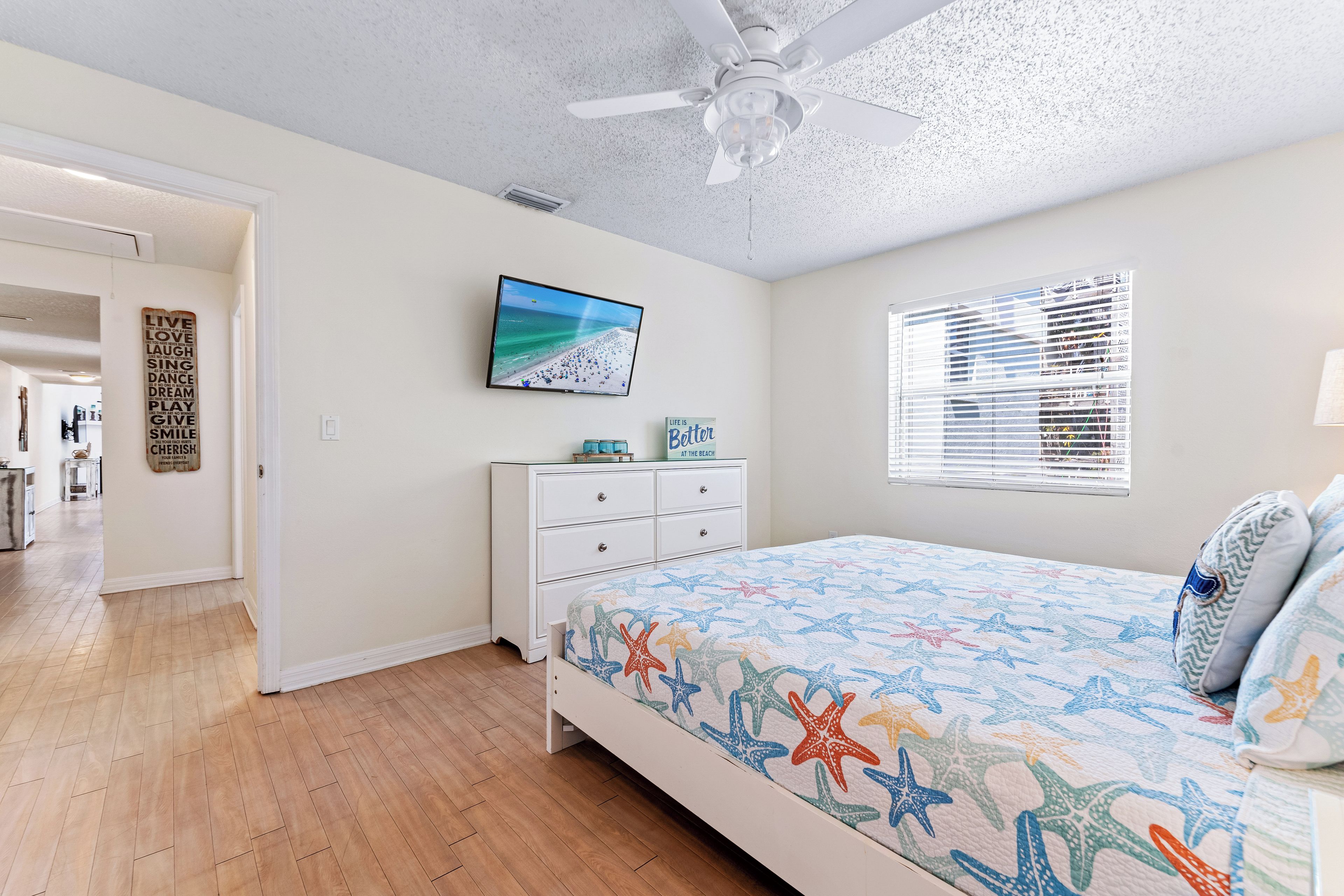 A bedroom with a bed, a ceiling fan, a TV on the wall, a window with blinds, and a hallway leading to another room.