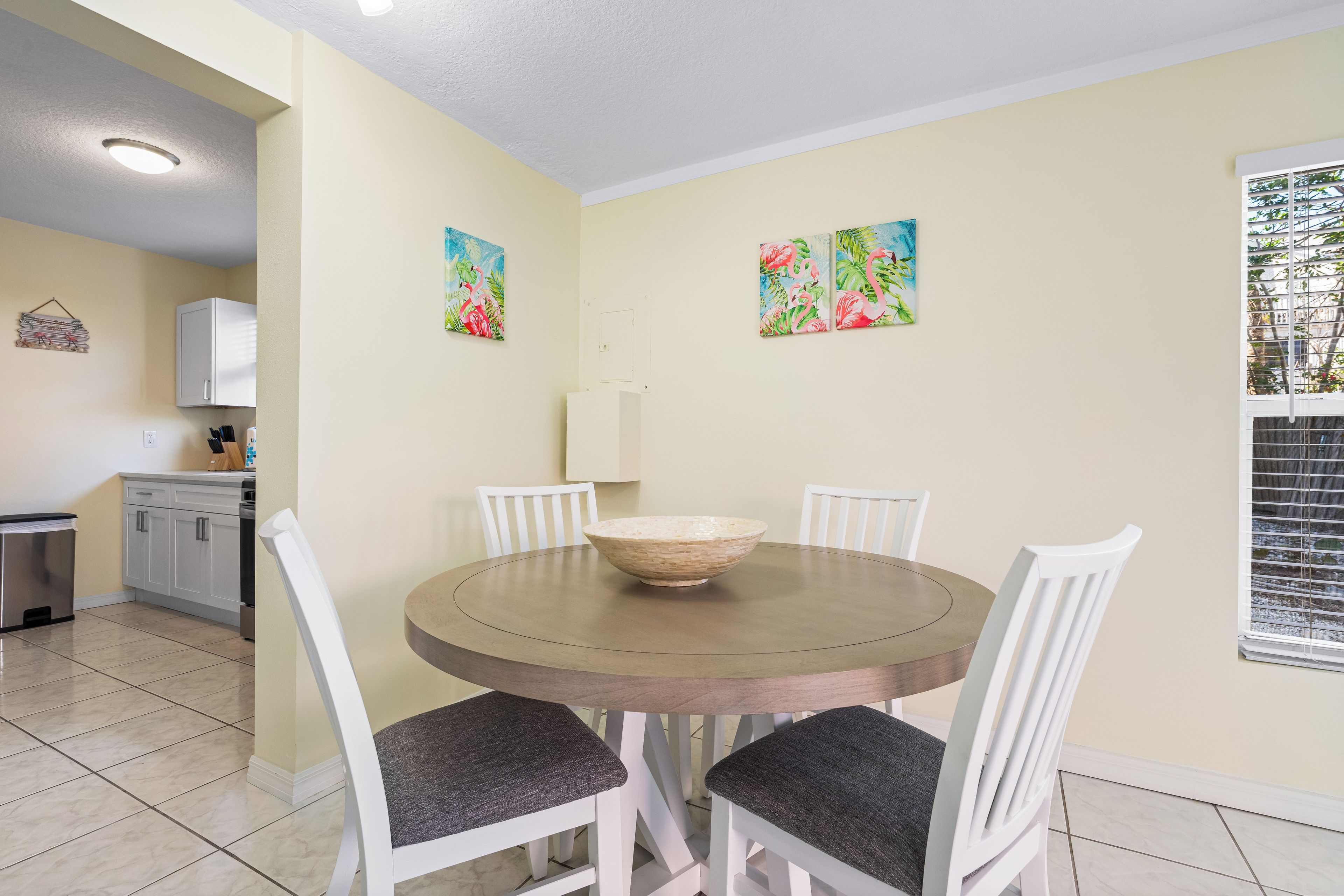 A dining area with a round table, white chairs, and a bowl. There are two wall-mounted paintings, a cabinet, and a window with blinds.