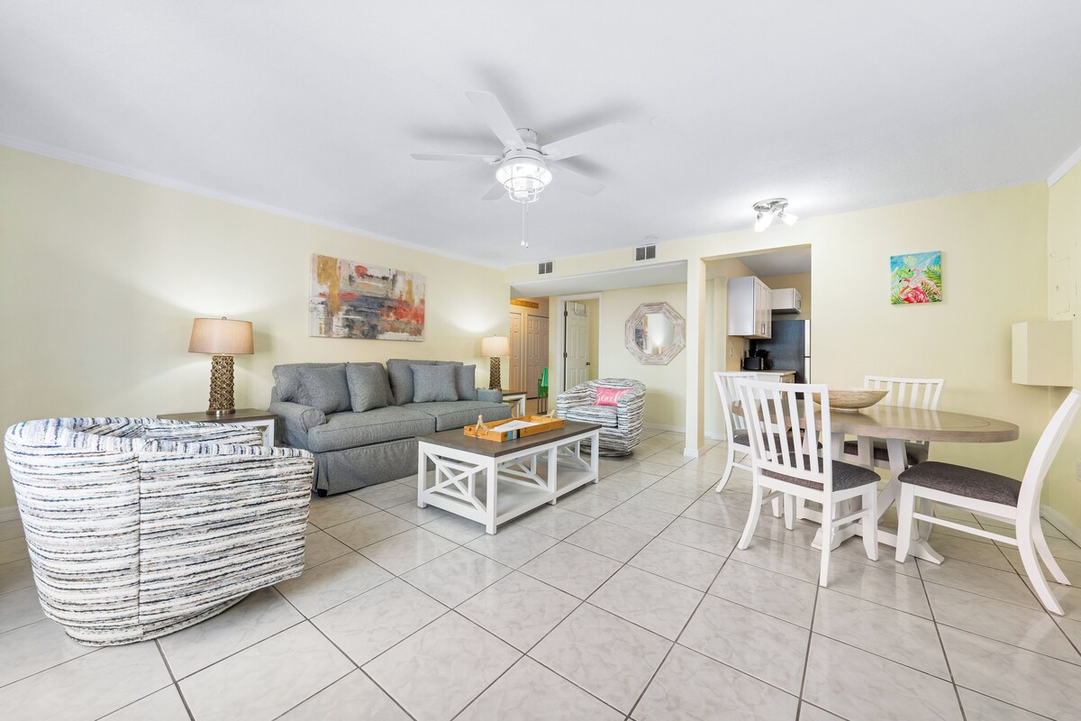 A living room with a sofa, coffee table, and dining table with chairs. There is a ceiling fan and a painting on the wall.