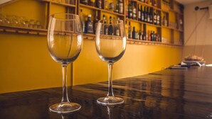 Two empty wine glasses on a bar counter with a yellow wall and shelves of liquor bottles in the background. - Restaurante Baia Guest House (São Vicente)
