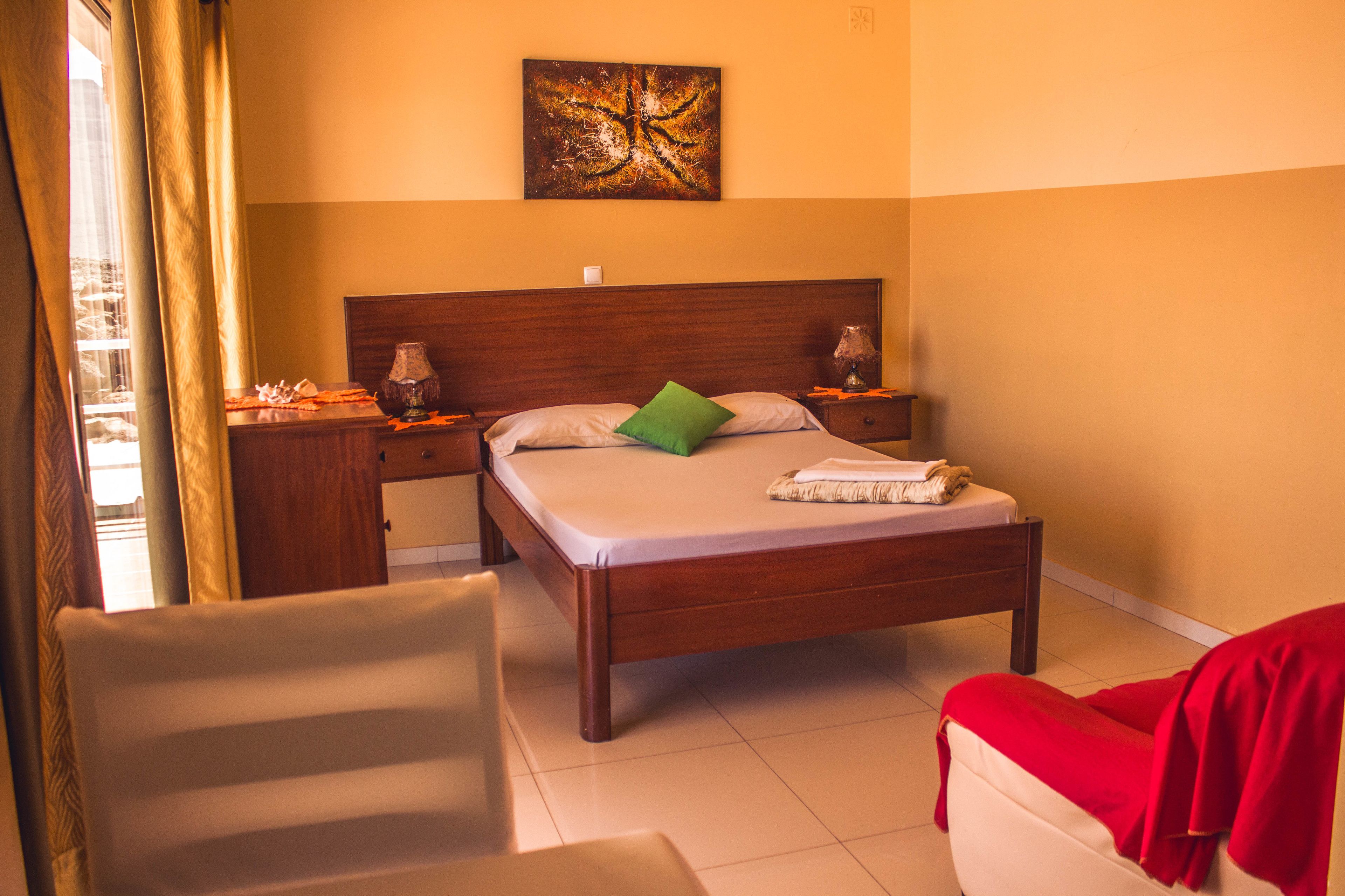 A hotel room with a wooden bed, a green pillow, a red blanket, a nightstand with a lamp, and a painting on the wall.