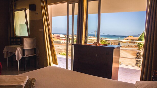 A hotel room with a bed, a desk, a chair, and a view of the ocean and beach. - Restaurante Baia Guest House (São Vicente)
