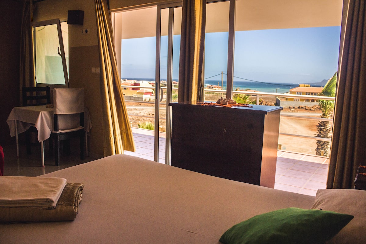 A hotel room with a bed, a desk, a chair, and a view of the ocean and beach.