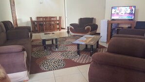 Ein Wohnzimmer mit einer Sofagarnitur, einem Couchtisch, einem gemusterten Teppich und einem Fernseher, der eine Nachrichtensendung zeigt.