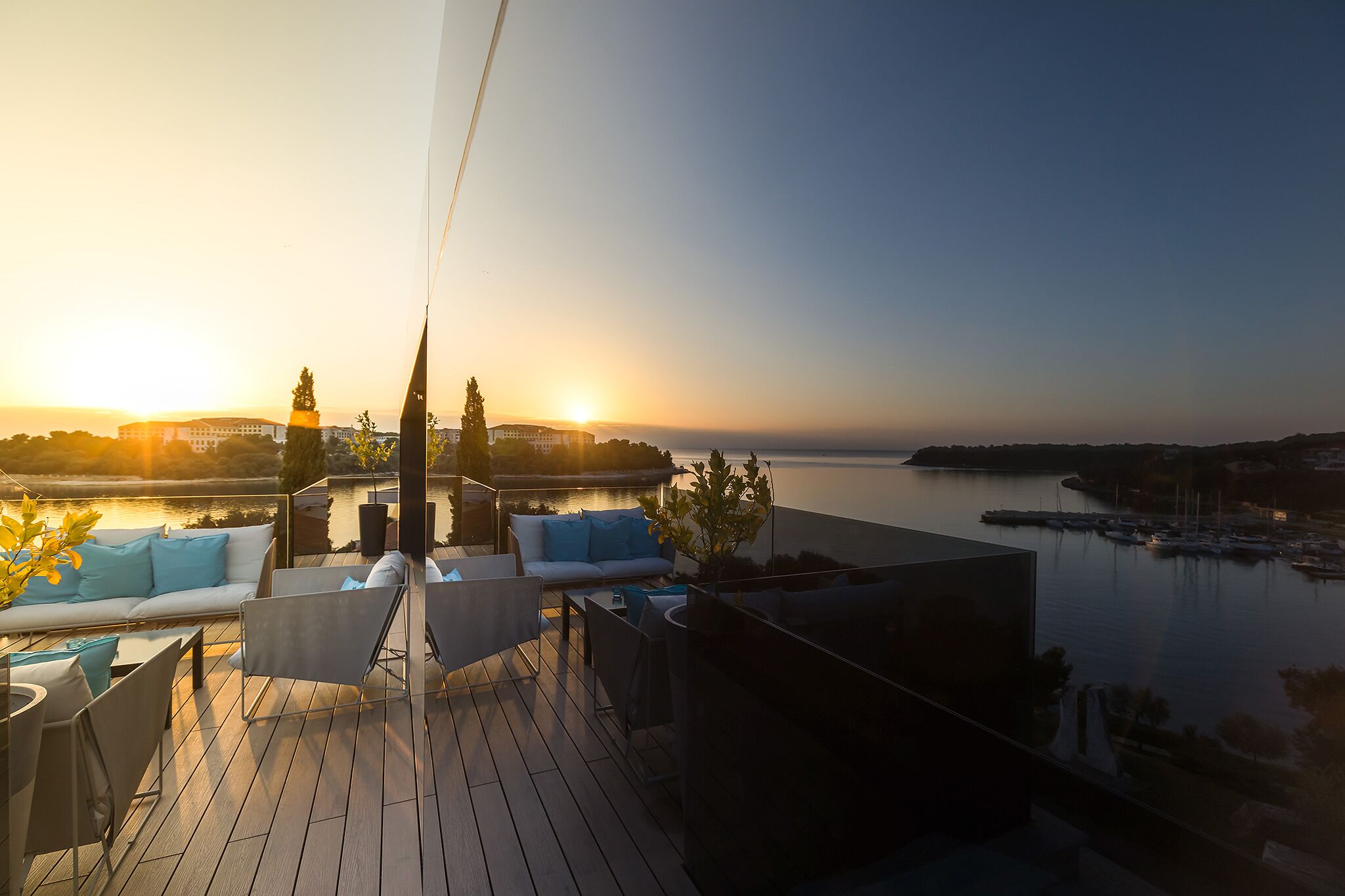 Terraza con mobiliario exterior, con vista a un cuerpo de agua y un edificio distante al atardecer.