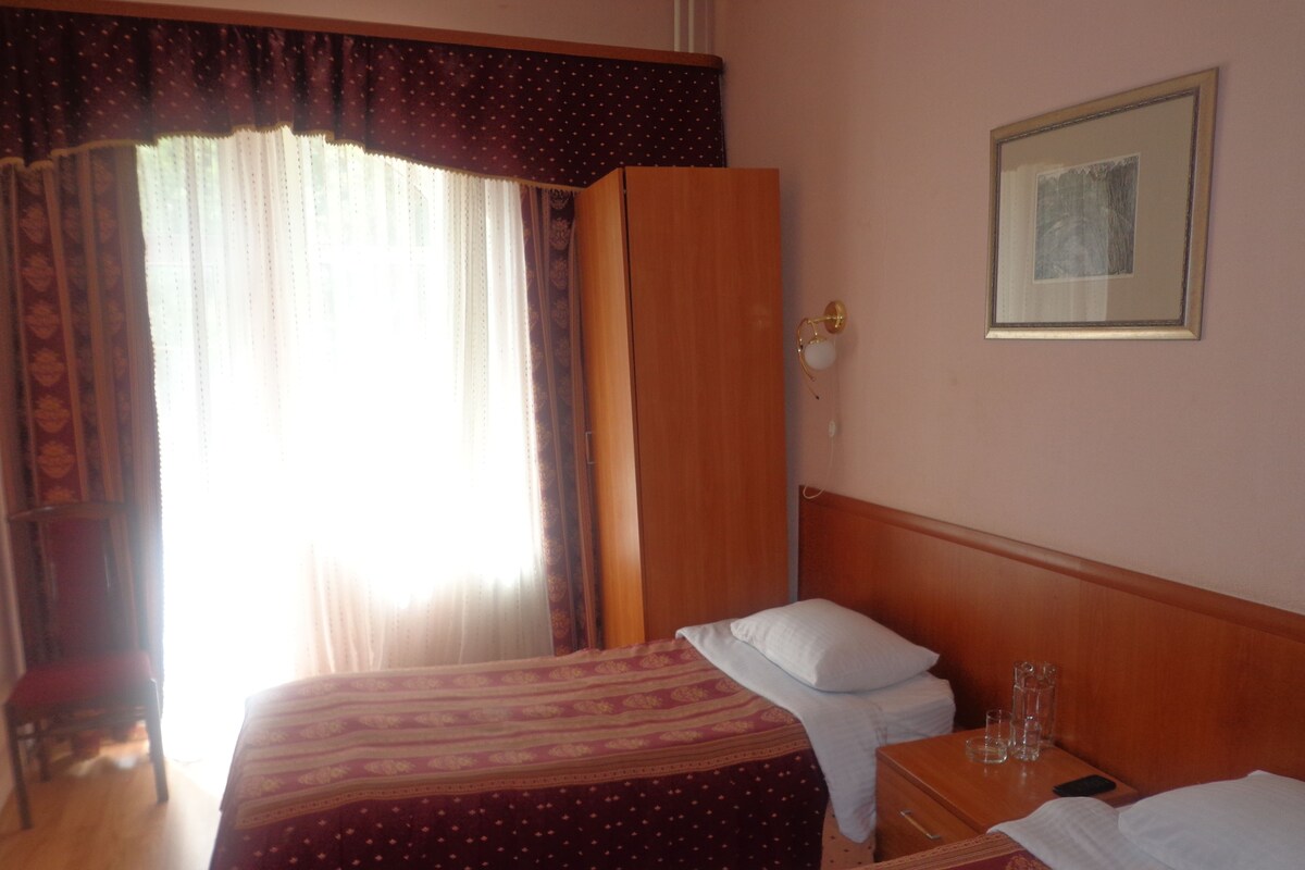 A hotel room with two beds, a wooden wardrobe, a bedside table with glasses, and a framed picture on the wall.