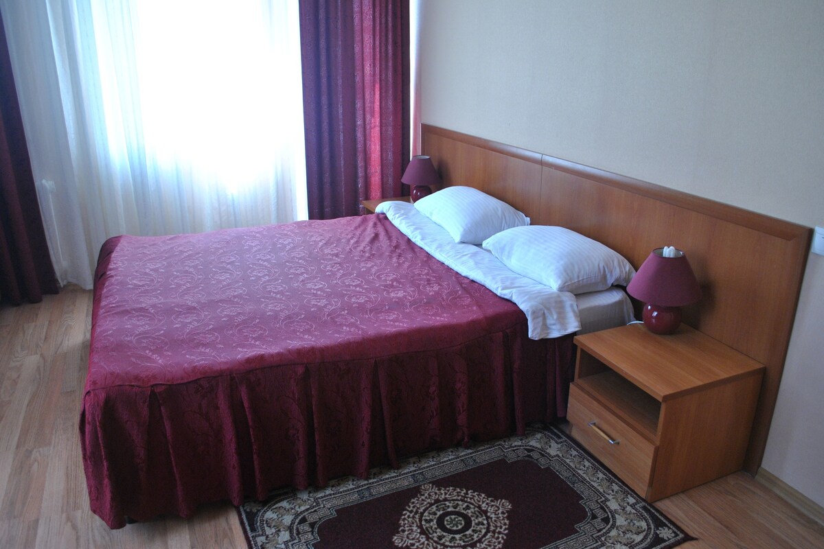 A neatly made bed with a red bedspread, two white pillows, and a wooden nightstand with a lamp.