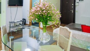 A dining area with a glass table, chairs, a vase of flowers, a TV mounted on the wall, and a door in the background. - Rustaveli Besiki Apartments (Tbilisi)