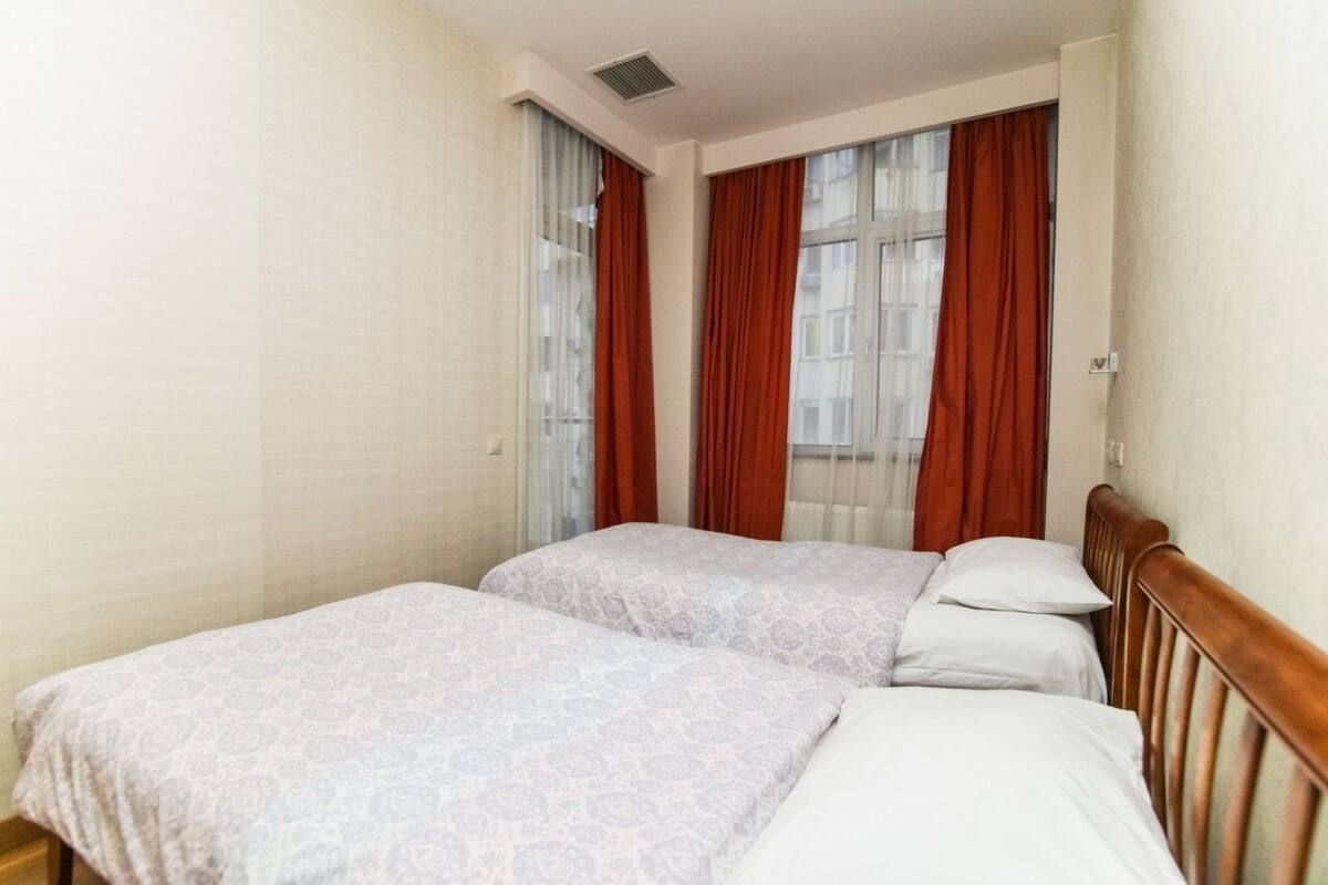 A hotel room with two single beds, a wooden headboard, a window with red curtains, and a light-colored wall.