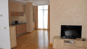 A living room with a flat-screen TV on a wooden stand, a kitchen area with wooden cabinets and a microwave, and a large window with curtains. - Rustaveli Besiki Apartments (Tbilisi)