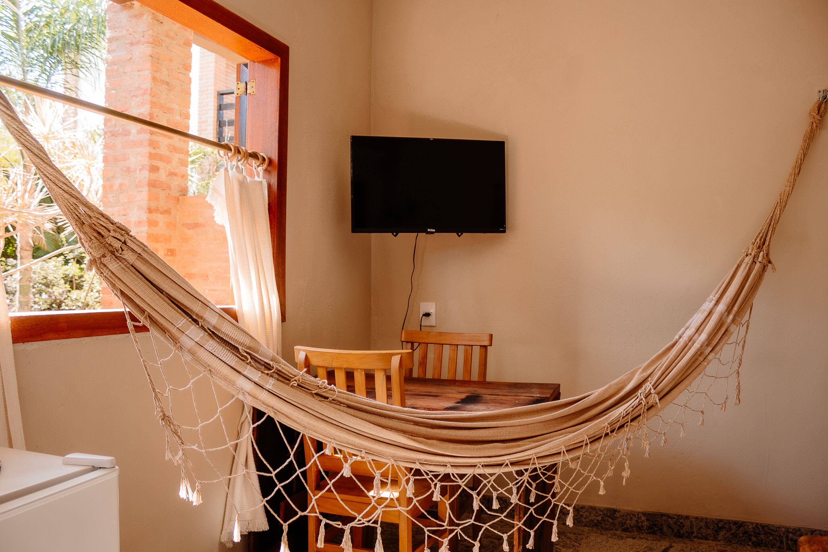 A hammock in a room with a TV mounted on the wall, wooden furniture, and a window with curtains.
