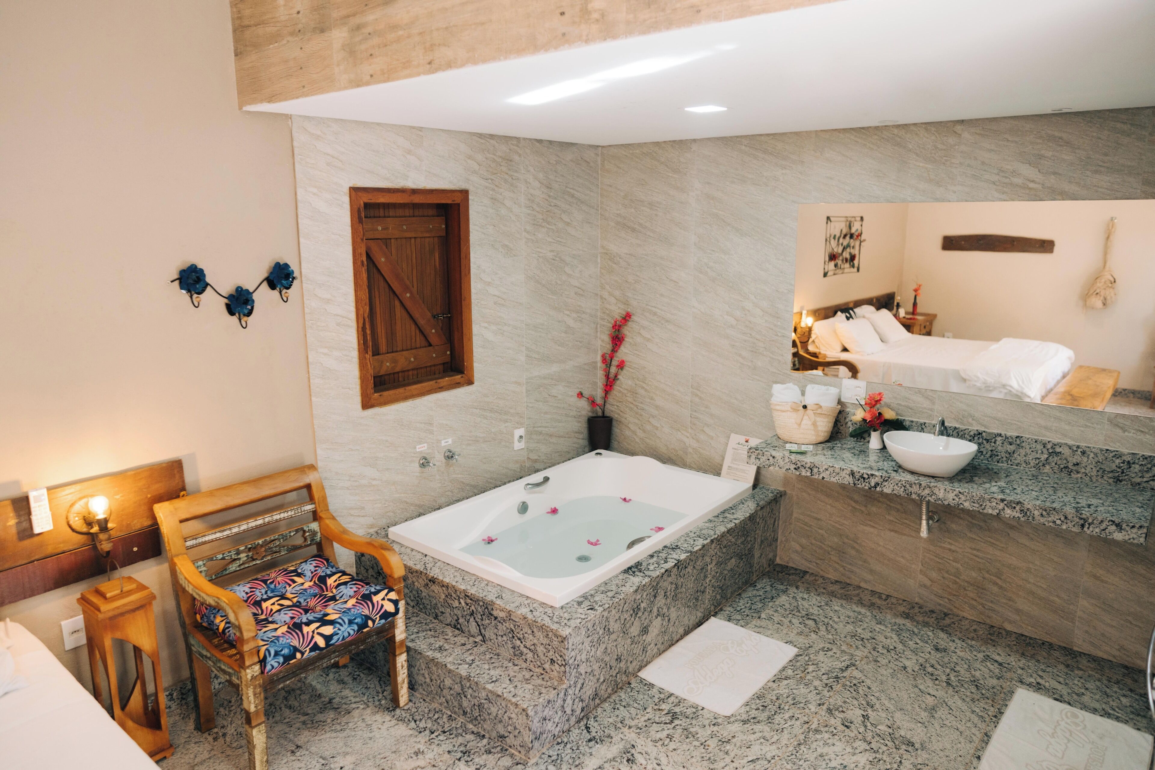A bathroom with a marble floor, a wooden chair, a bathtub, a vanity with a mirror, and a bed visible in the reflection.