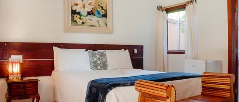 A hotel room with a bed, a bedside table with a lamp, a wooden bench, a painting on the wall, and a window with curtains.