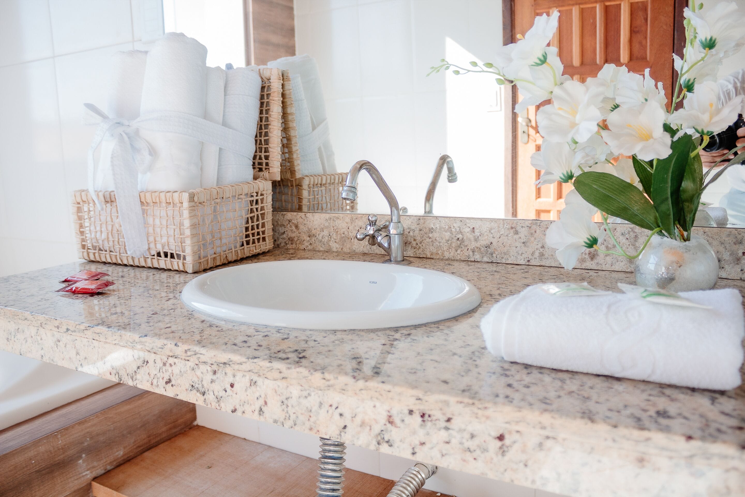 A bathroom with a granite countertop, a white sink, and a mirror. There are towels and a basket of toiletries.
