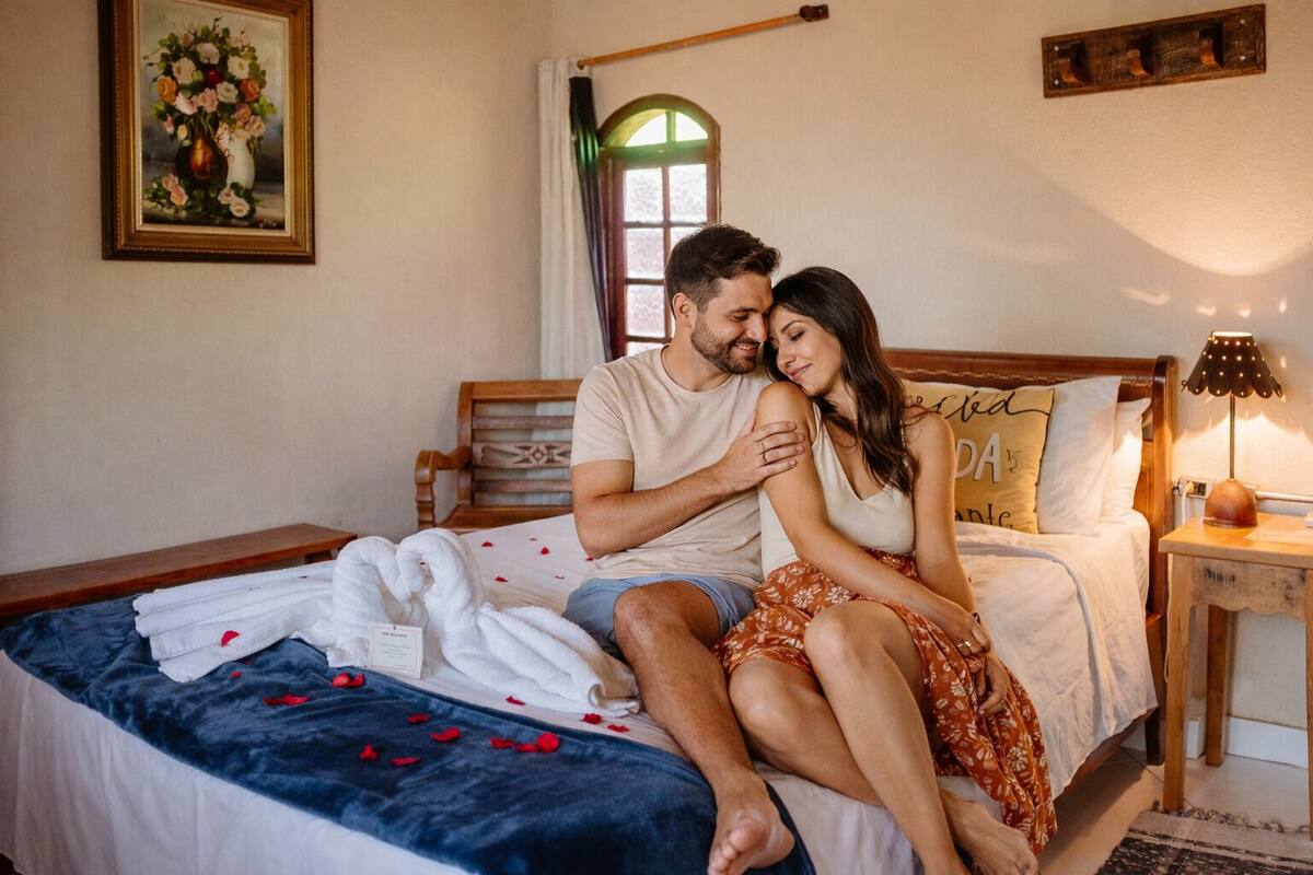A couple sitting on a bed with a blue blanket and white towels, a wooden headboard, a bedside lamp, and a painting on the wall.