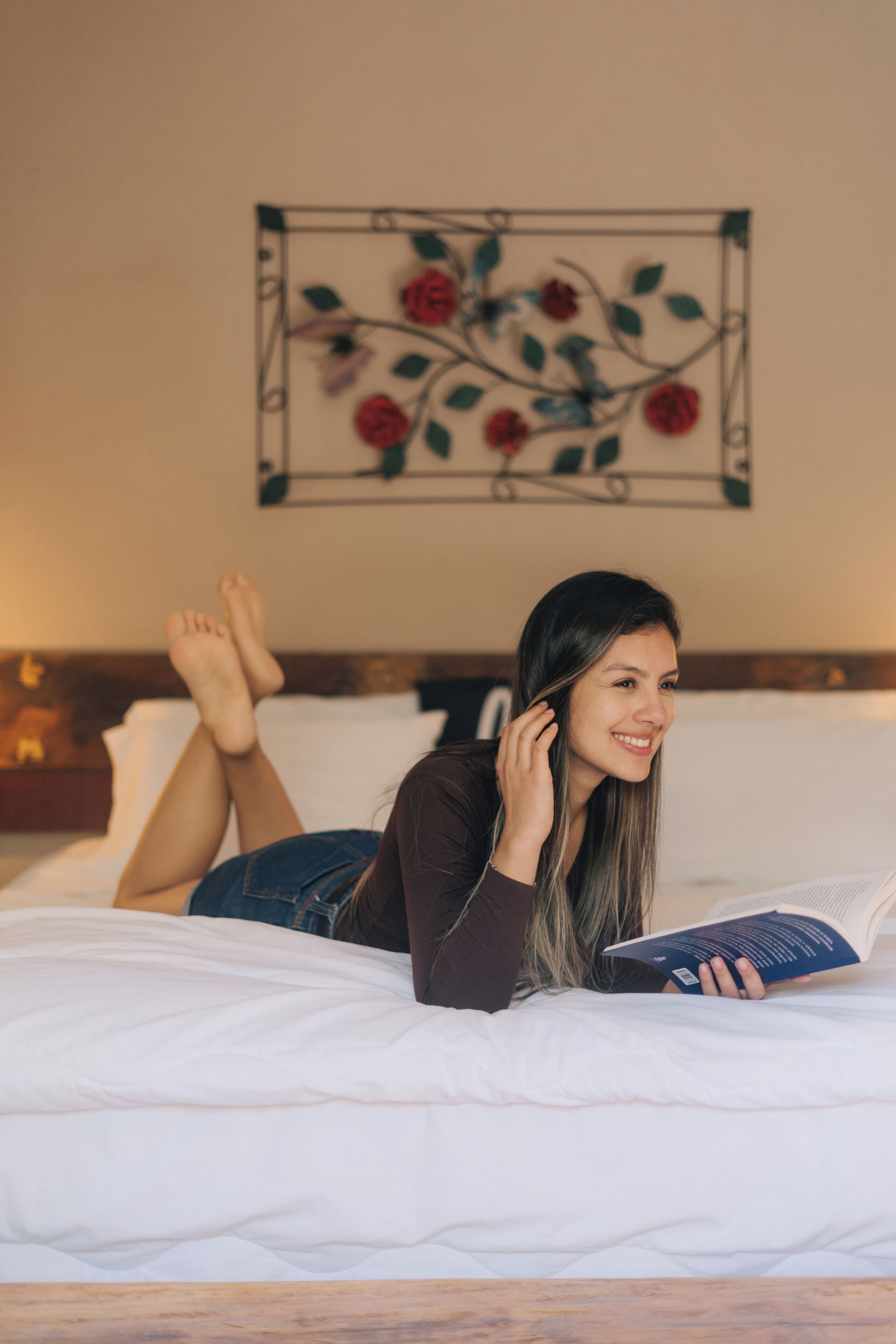 A person lying on a bed with a book, in a room with a floral wall decoration.