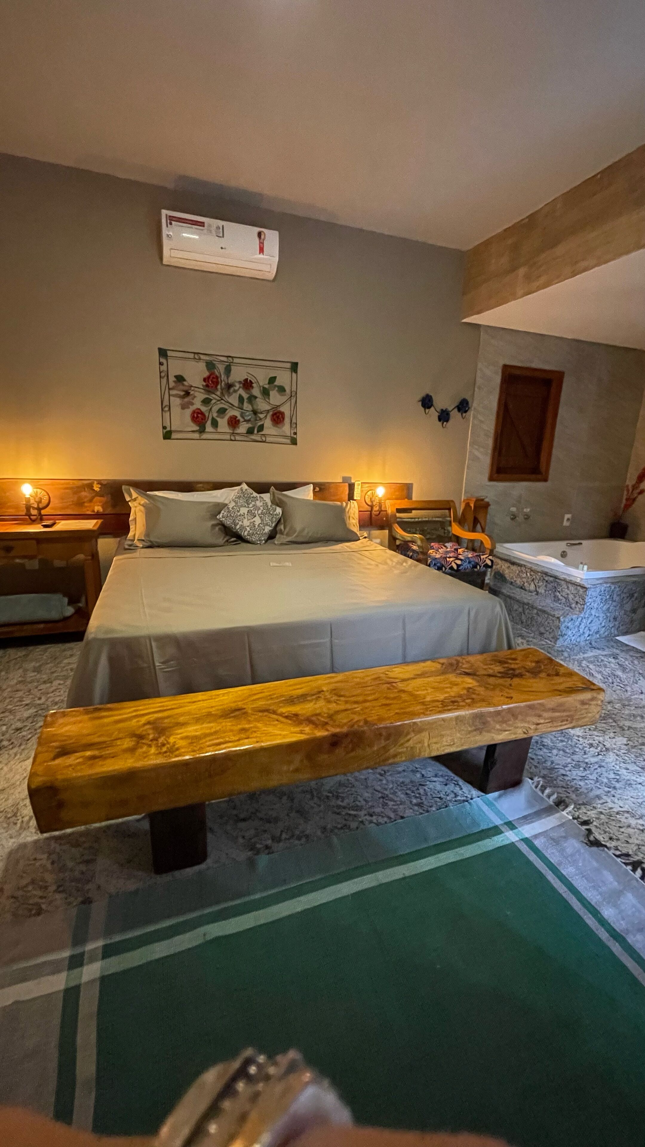 A bedroom with a large bed, a wooden bench, a green area rug, a wall-mounted air conditioning unit, and a bathtub.