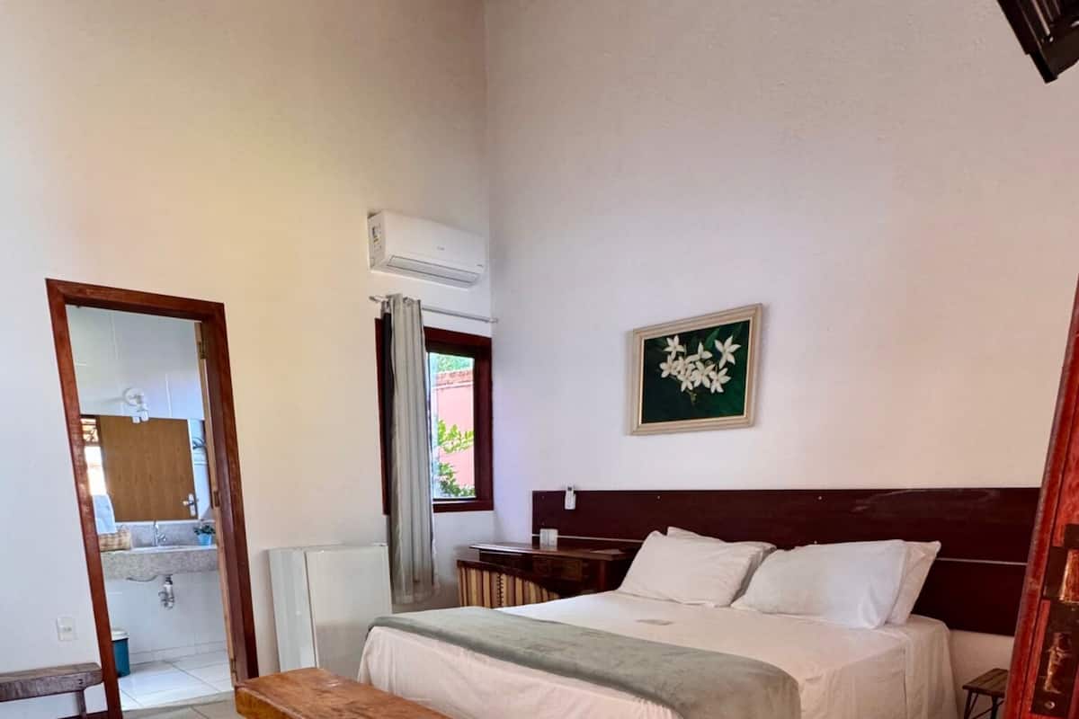 A bedroom with a wooden bench, a bed with white bedding, a wall-mounted air conditioner, and a framed picture.