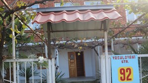 Un bâtiment doté d’une entrée couverte, d’une enseigne indiquant « rue Vachlovani, 92 », et d’une entrée fermée par un portail, avec un chemin menant à la porte.