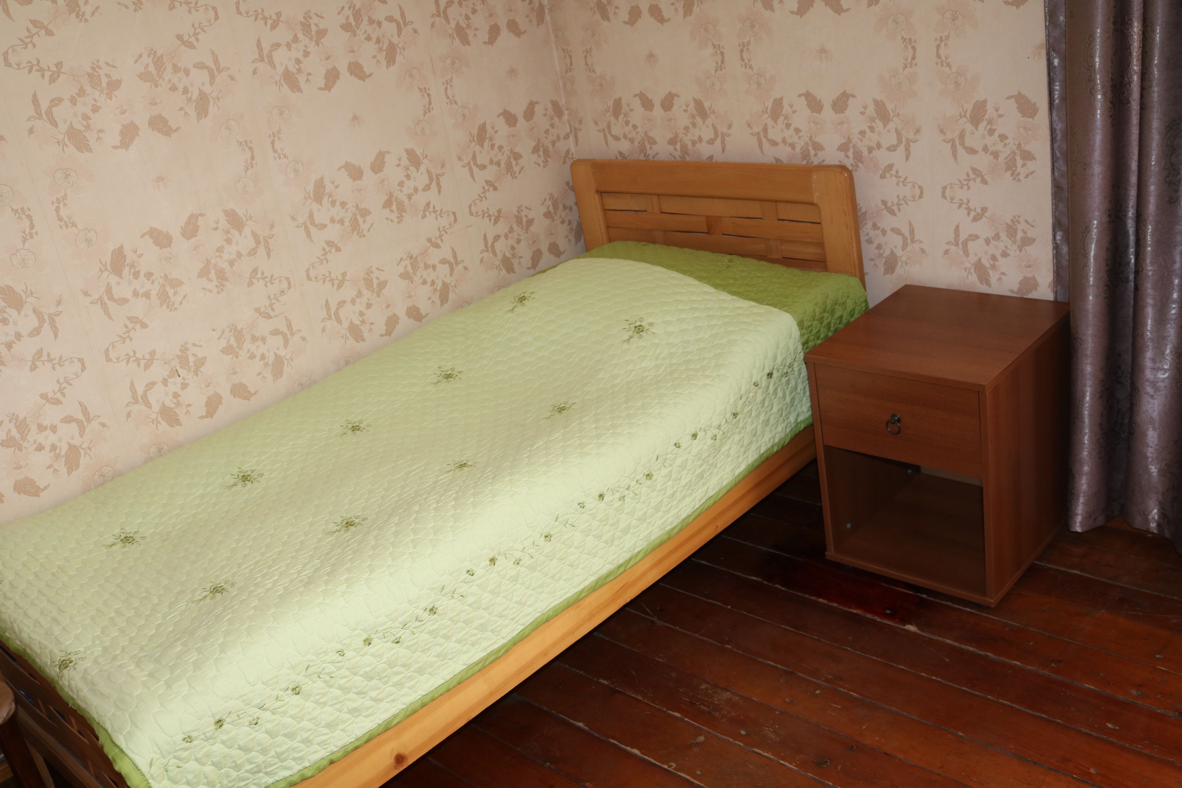 A single bed with a wooden headboard and a green quilt, a wooden nightstand, and a wooden wardrobe.