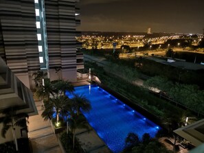 A nighttime view of a hotel with a swimming pool, illuminated by blue lights, surrounded by greenery and a distant cityscape. - Cozy Condo at The Meridin Medini (Iskandar Puteri)