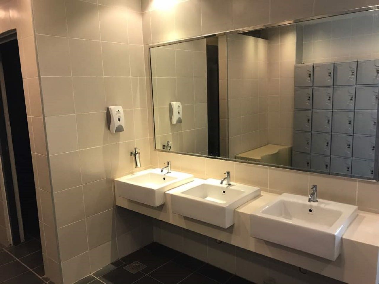 A modern bathroom with three sinks, a large mirror, and a row of lockers.