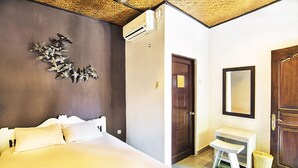 A bedroom with a bed, a small bedside table, a mirror, and an air conditioning unit. - Puri Agung Homestay (Kuta)
