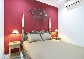 A bedroom with a red wall, a bed with a wooden headboard, two bedside tables with lamps, and an air conditioning unit on the wall.