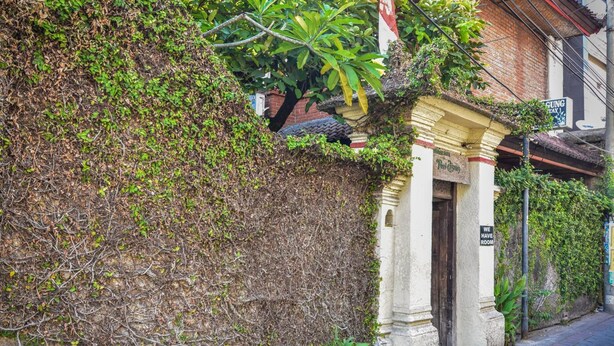 A building with a partially covered entrance, a sign indicating rooms are available, and a brick wall with overgrown vegetation. - Puri Agung Homestay (Kuta)