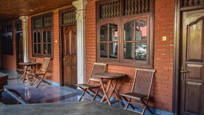 A covered outdoor seating area with wooden chairs and tables, brick walls, and large wooden doors. - Puri Agung Homestay (Kuta)
