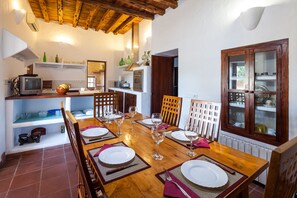 On aperçoit un coin repas avec une table en bois dressée pour six personnes, entourée de chaises. Au fond, on distingue une cuisine équipée d’une cuisinière, d’un four et d’étagères.