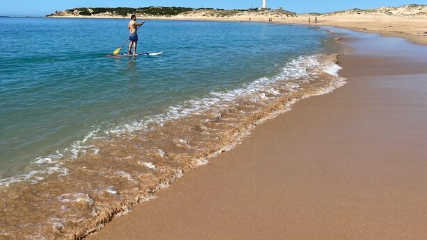 Una playa con aguas azul cristalino, una orilla arenosa y un faro a lo lejos.