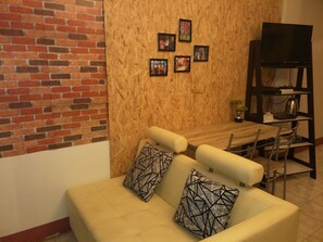 A living room with a brick wall, a wooden wall with framed pictures, a beige sofa with patterned cushions, a wooden dining table with chairs, and a TV on a stand.