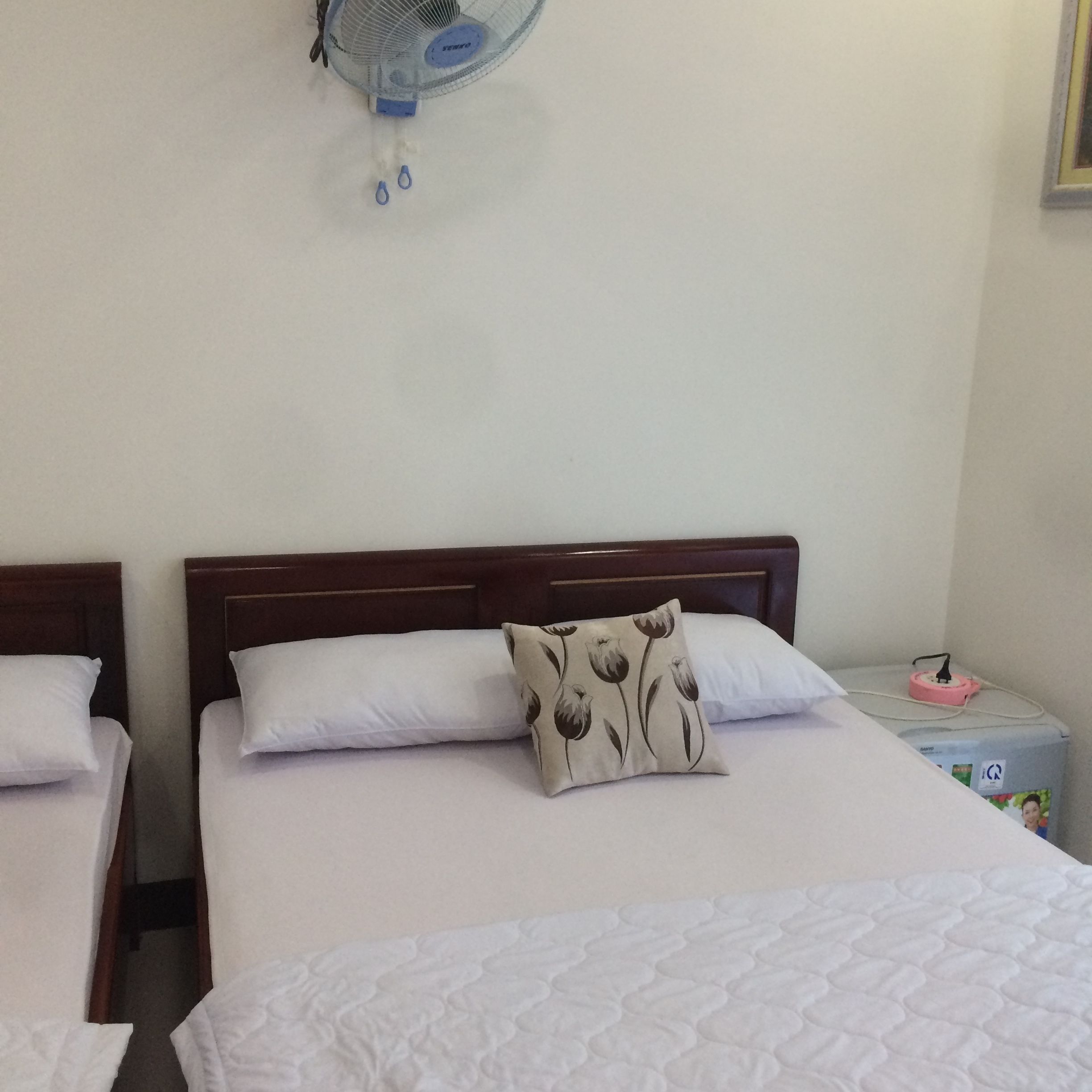 Habitación de hotel con dos camas individuales, cada una con ropa de cama blanca y cabeceros de madera. Se observa un ventilador de pared sobre las camas.