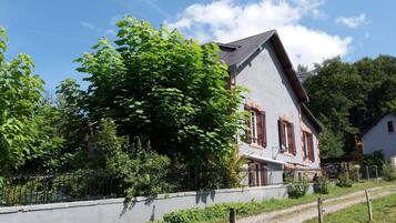 Une maison à deux étages avec un toit en pente, un grand arbre à l’avant et un chemin de gravier menant à l’entrée.