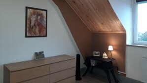 Une pièce avec un plafond en pente, une commode en bois, un tableau encadré, une petite table surmontée d’une statue et une fenêtre laissant entrer la lumière naturelle.