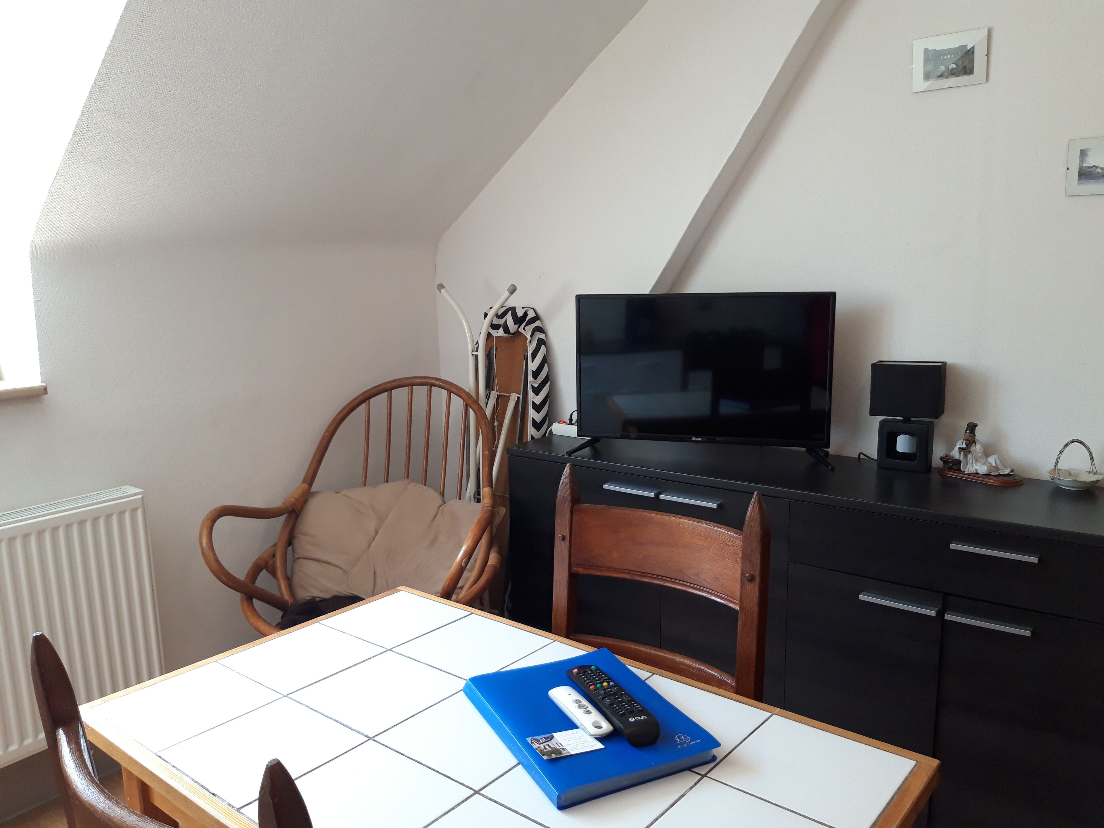 Une pièce comprenant un téléviseur posé sur un meuble, une chaise, une table avec une télécommande et un radiateur.