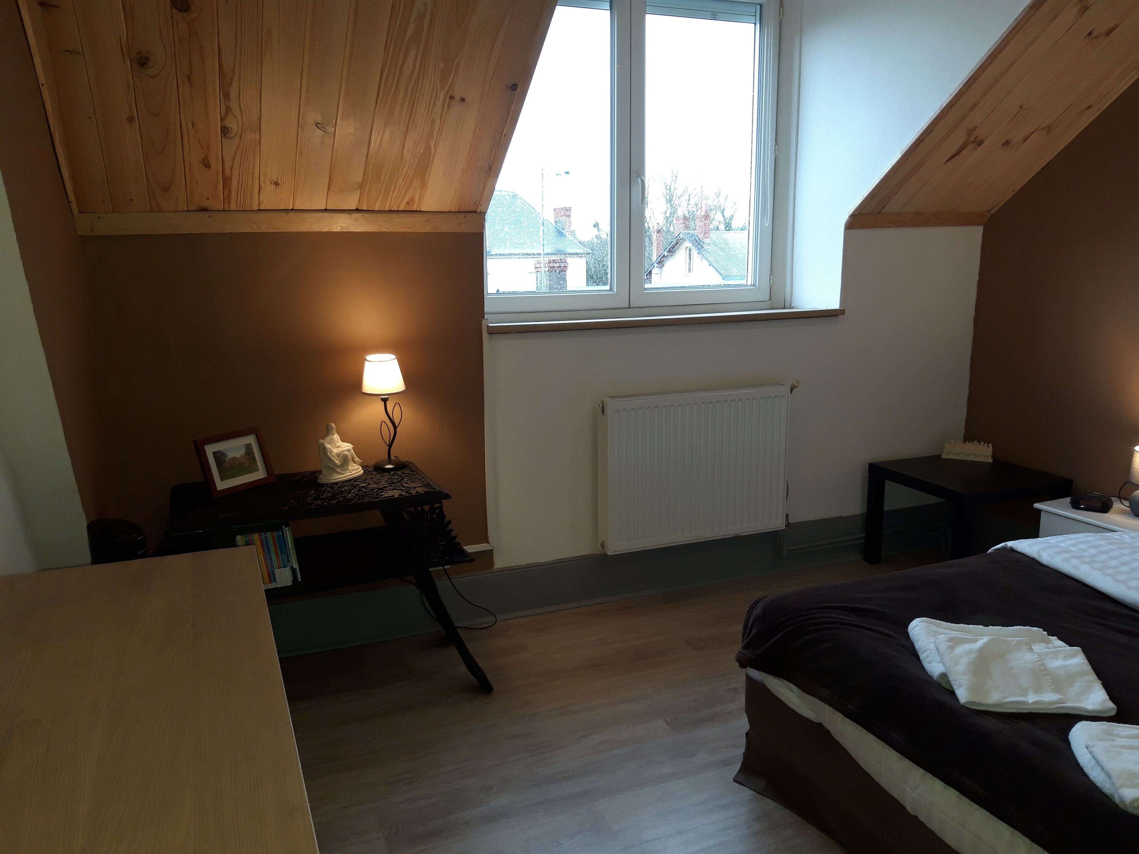 Ett sovrum med ett sluttande tak, en säng med vita sängkläder, ett skrivbord med en lampa och ett fönster med utsikt över byggnaderna utanför.