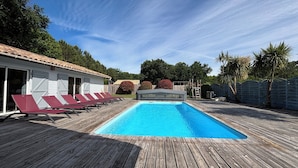 A wooden deck with a swimming pool, red lounge chairs, and a building with a tiled roof. - Near Soulac-sur-Mer, 6-bedroom villa with heated indoor pool, jacuzzi, and sauna (Grayan-Et-L'hôpital)