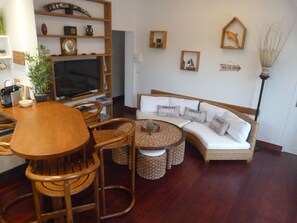 Un salon comprenant une table à manger en bois, des chaises en osier, un canapé blanc avec des coussins, un téléviseur et une bibliothèque agrémentée d’objets décoratifs.