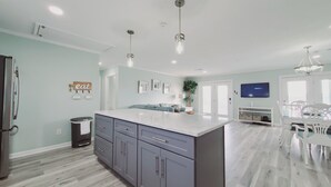 A modern kitchen with grey cabinets, a large island, pendant lighting, and a flat-screen TV mounted on the wall.