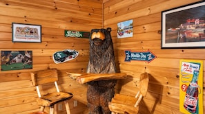 Interior de una cabaña de madera con una escultura de oso, sillas de madera y varios carteles, incluyendo un anuncio de Pepsi Cola.