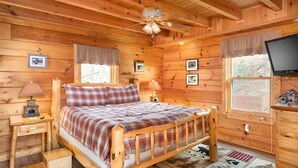 A cozy log cabin bedroom with a wooden bed, a nightstand, a lamp, a ceiling fan, and a TV on a wooden stand.