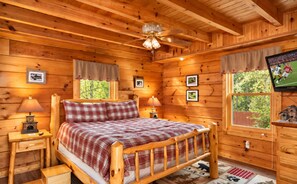 A wooden cabin bedroom with a bed, two bedside tables, a ceiling fan, and a television on the wall.