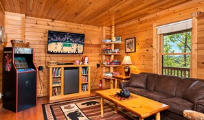 A cozy cabin interior with a basketball arcade game, a bookshelf, a couch, and a window with a view of trees.