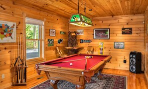 A pool table with a red felt surface, surrounded by wooden walls and a hanging light fixture.