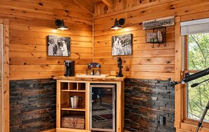 Interior de una cabaña de madera con una estación de café, dos lámparas de pared, una cafetera, un hervidor, una botellera y un telescopio.