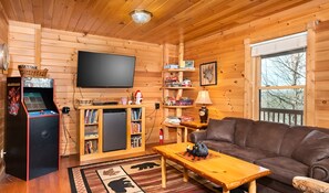 Tunnelmallinen mökin sisustus, jossa on puusisustus, seinälle asennettu televisio, pieni jääkaappi, kirjahylly, sohva ja sohvapöytä.