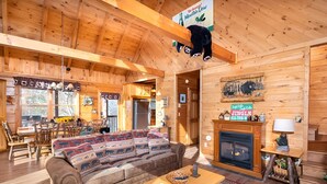 A cozy cabin interior with a wooden ceiling, a fireplace, a sofa with patterned cushions, and a dining area with a table and chairs.